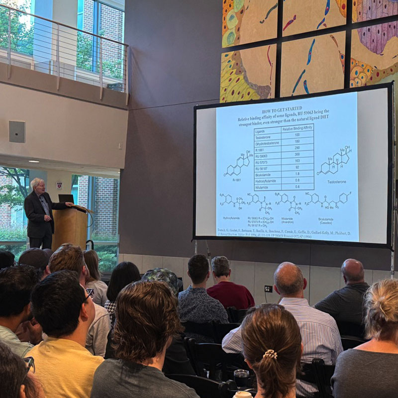 A speaker presents chemical diagrams and data on a large projection screen to an audience seated in a lecture hall.