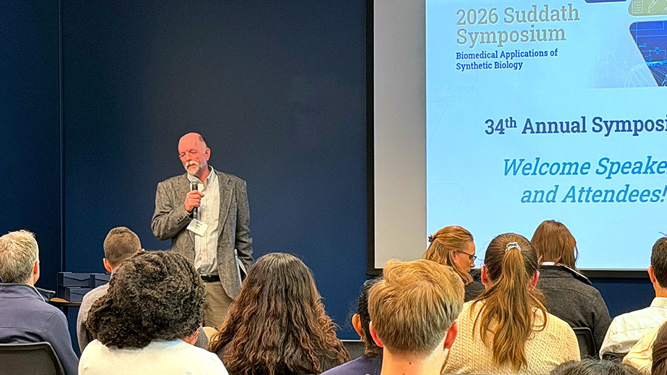 A man in a gray suit jacket speaks into a microphone in front of a crowd of people in a seminar room. A slide in the background says "2026 Suddath Symposium".