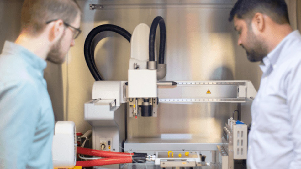 Two researchers observing a 3D bioprinting system in a laboratory, with a bioprinter positioned inside a protective enclosure.